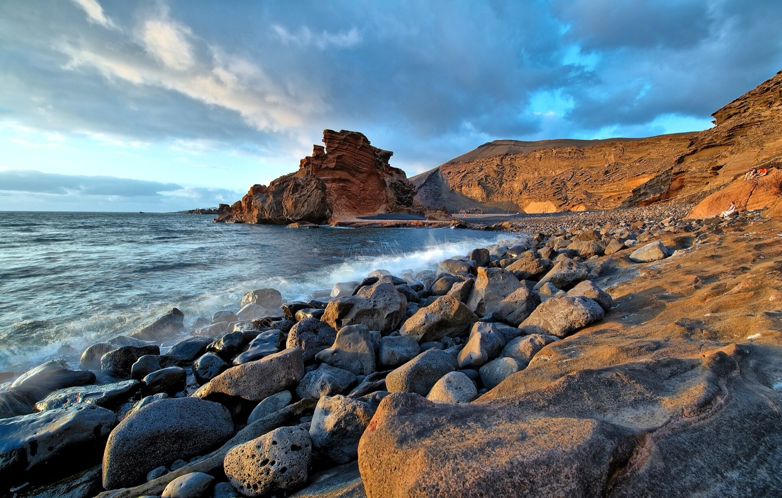 Costa de El Golfo al atardecer: cantos rodados volcánicos, olas blancas y acantilados rojizos bajo un cielo de nubes.
