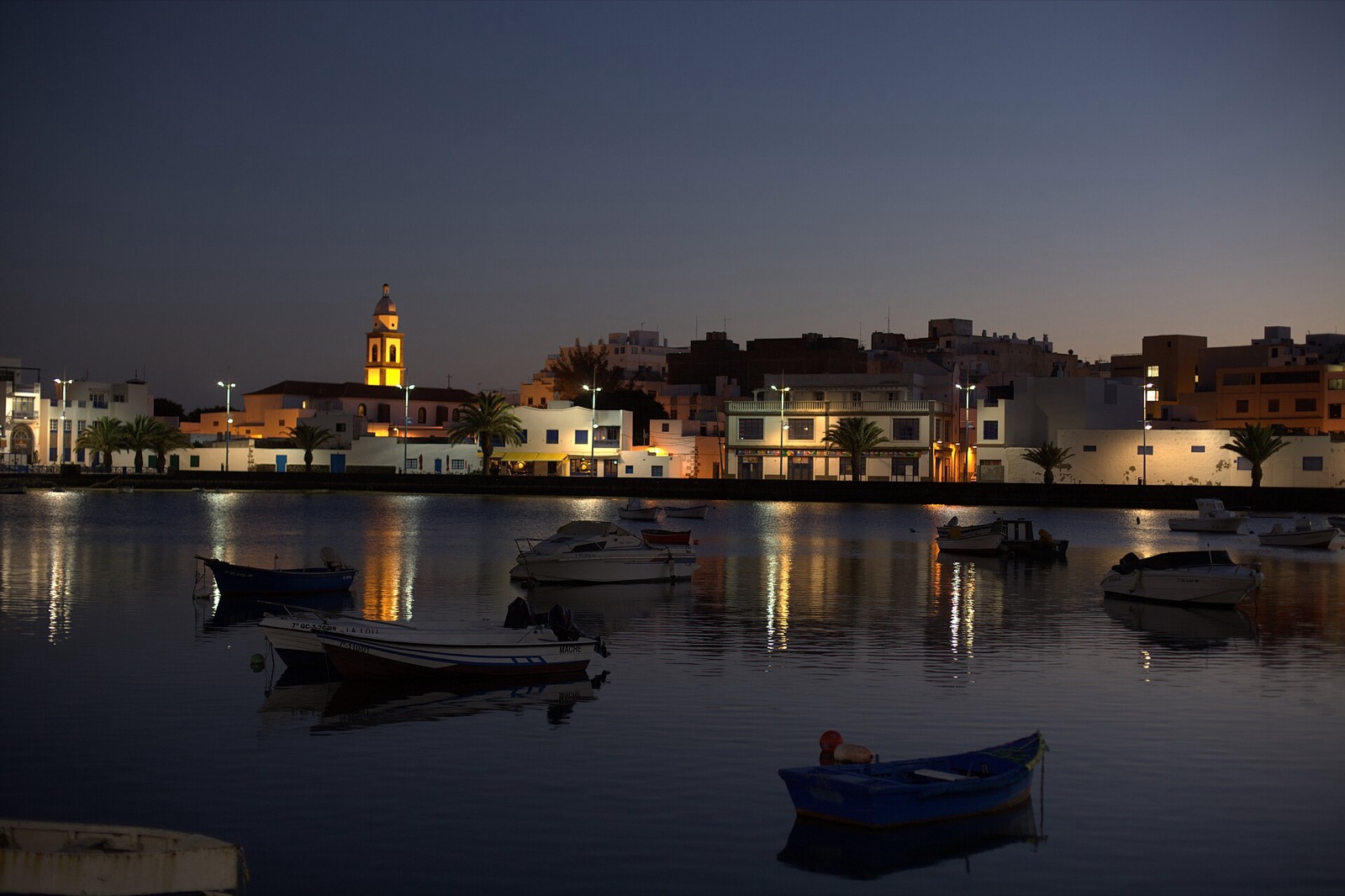 El Charco de San Ginés al anochecer: barcas quietas sobre el agua y, al fondo, las luces del paseo y el campanario de la iglesia.