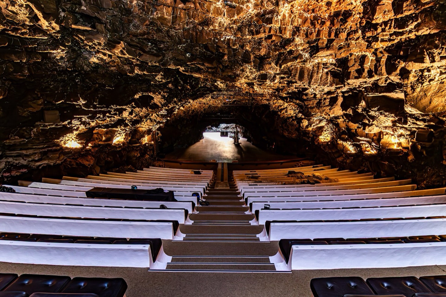 El auditorio de Jameos del Agua: filas de asientos blancos bajo la bóveda de lava del tubo volcánico, iluminada en tonos cálidos.