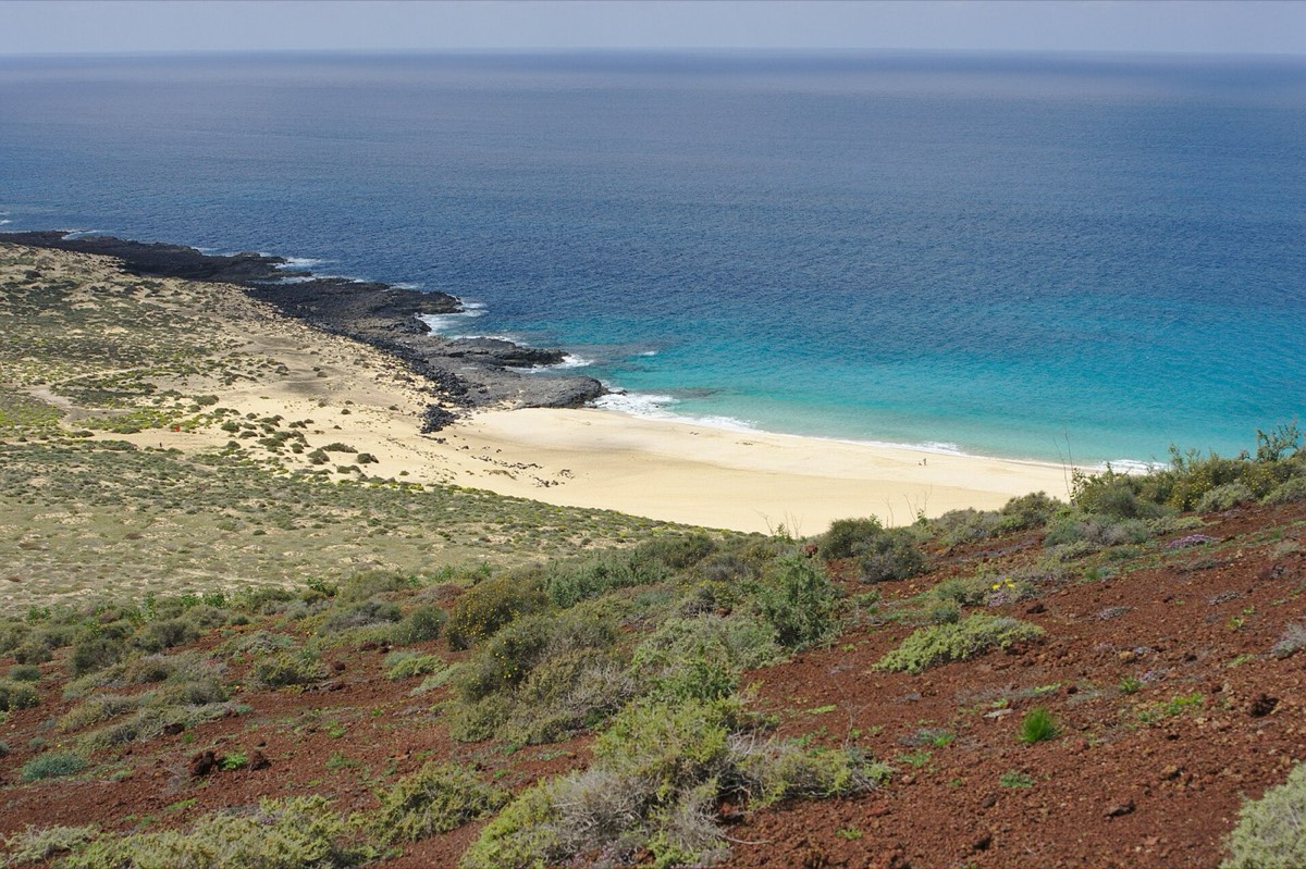 Playa de Las Conchas vista desde la ladera rojiza de Montaña Bermeja, con el mar turquesa